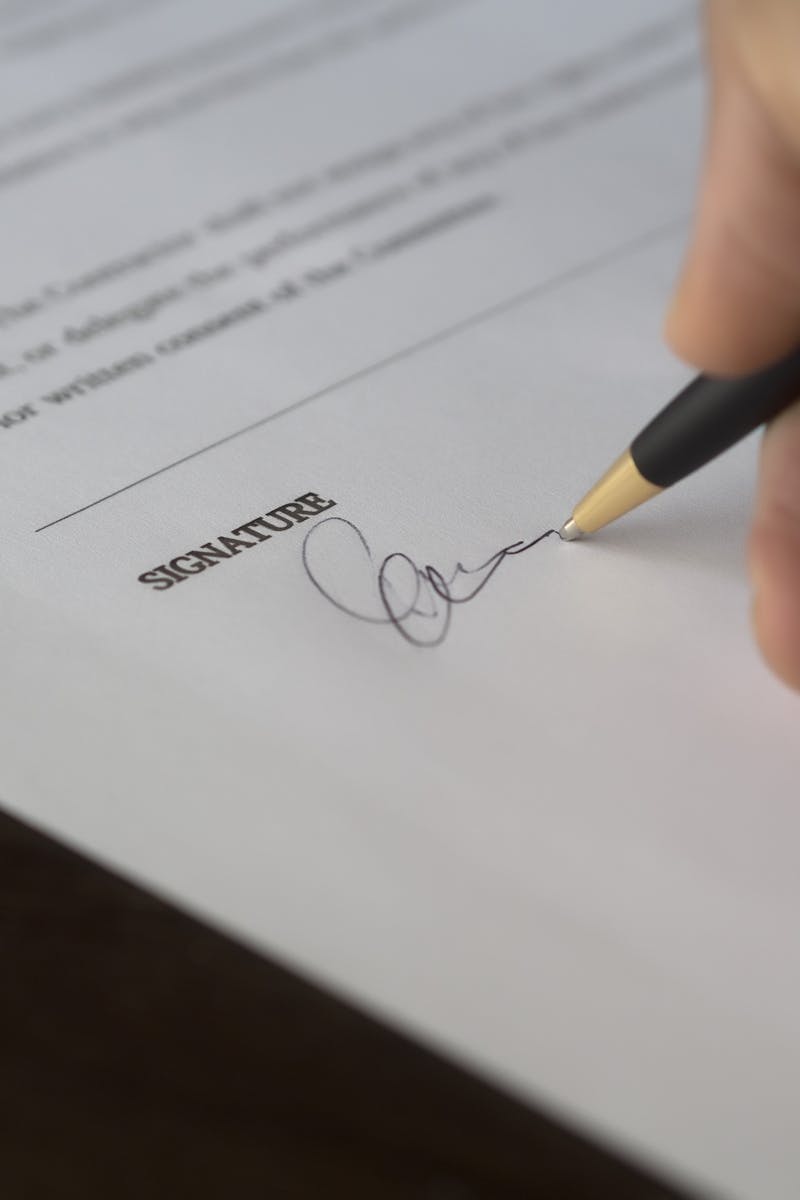 A hand signs a formal contract with a pen on a wooden desk.